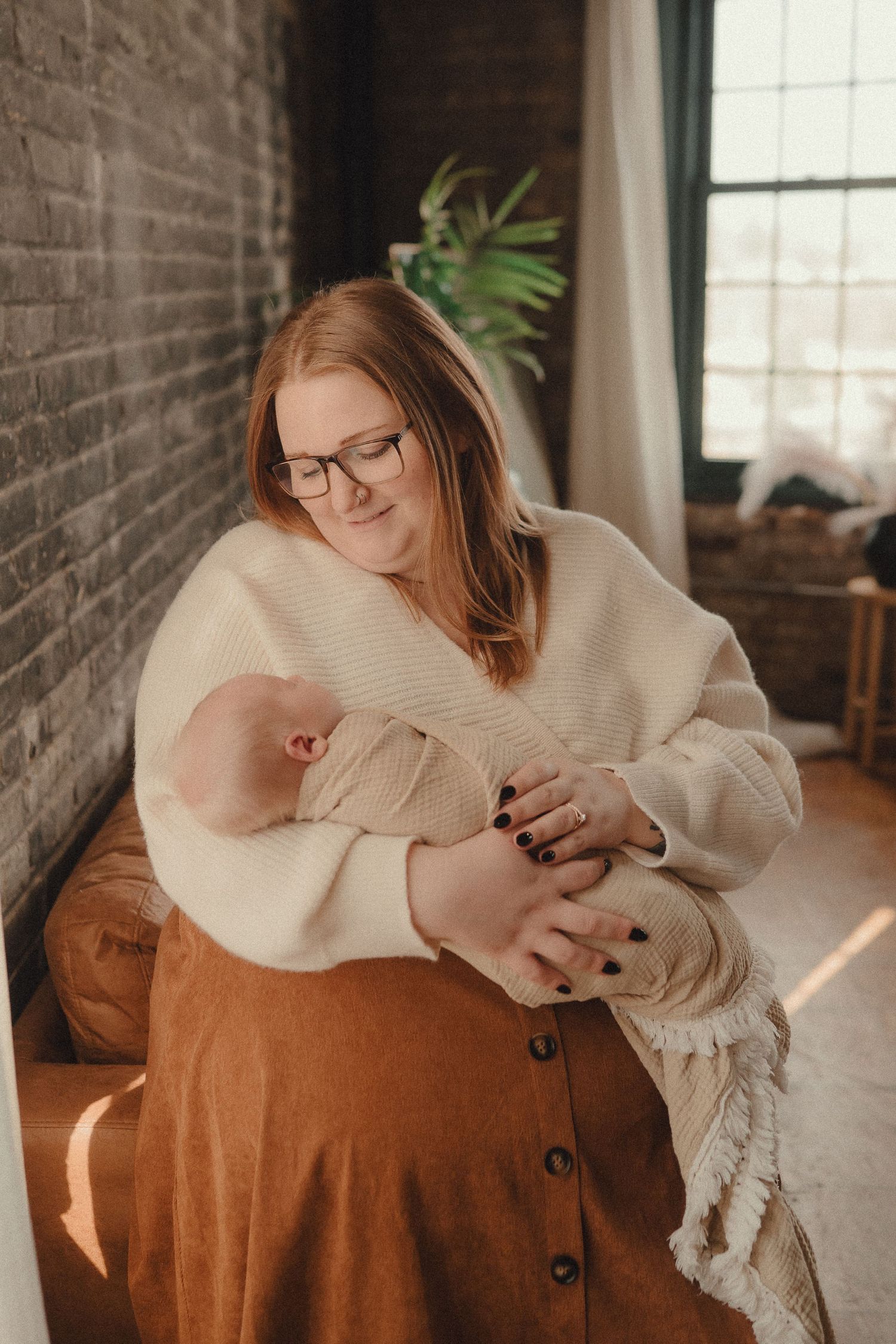 A red-haired mother wearing glasses and a cream sweater tenderly holds her newborn baby in a sunlit room.