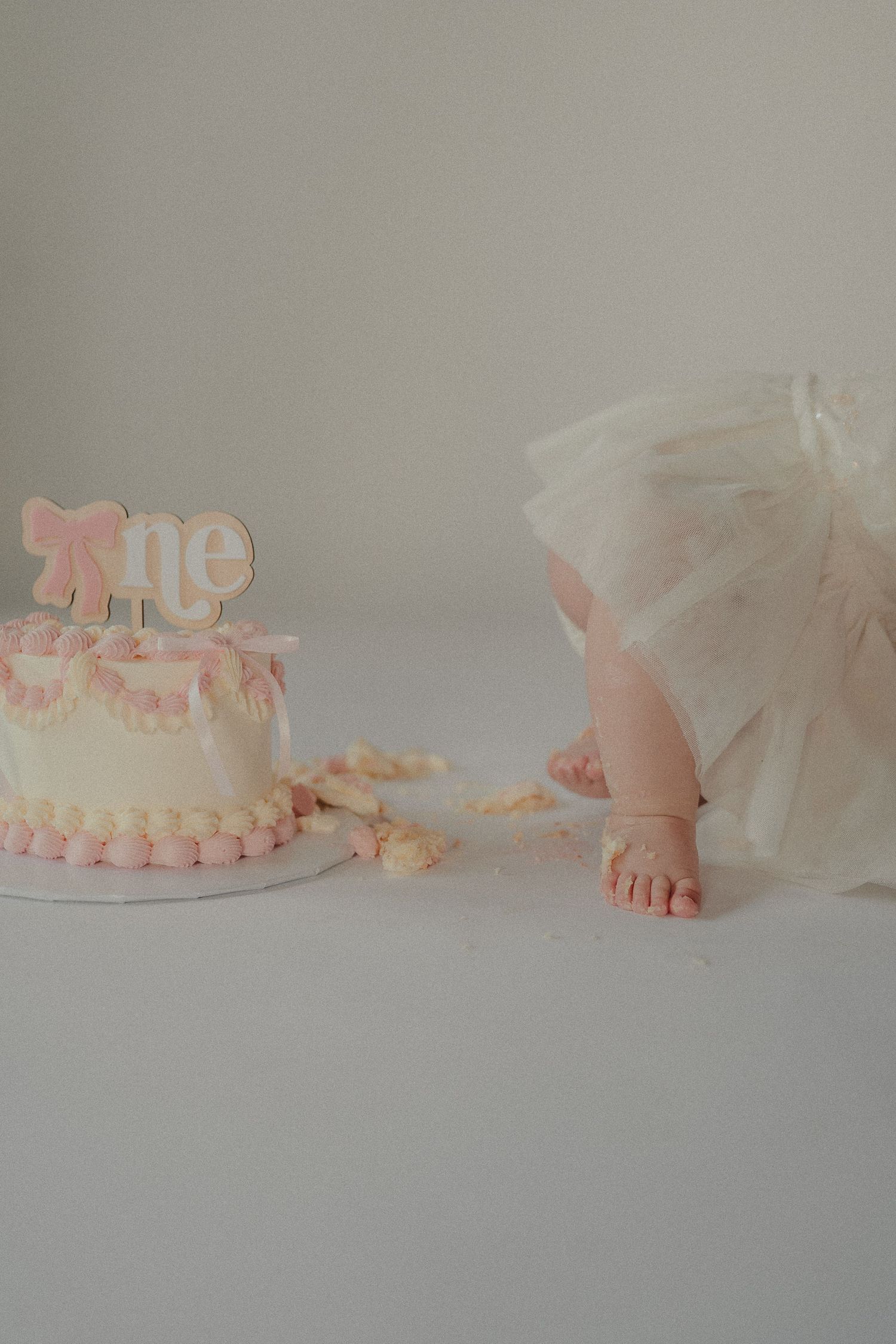 Close-up of baby feet next to a pink and white first birthday smash cake with bow topper.