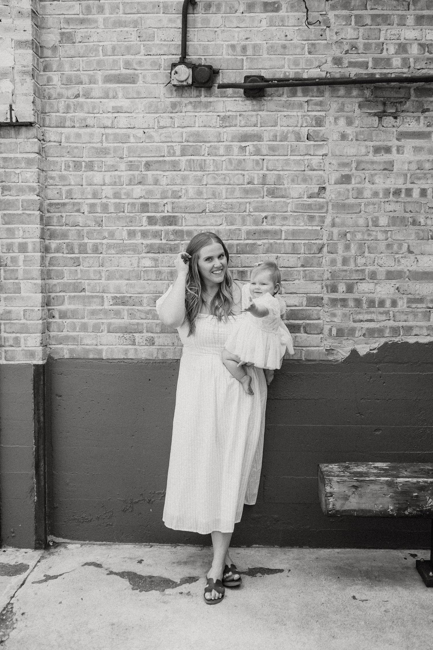 Black and white portrait of a mother in white dress holding her baby girl against brick wall.