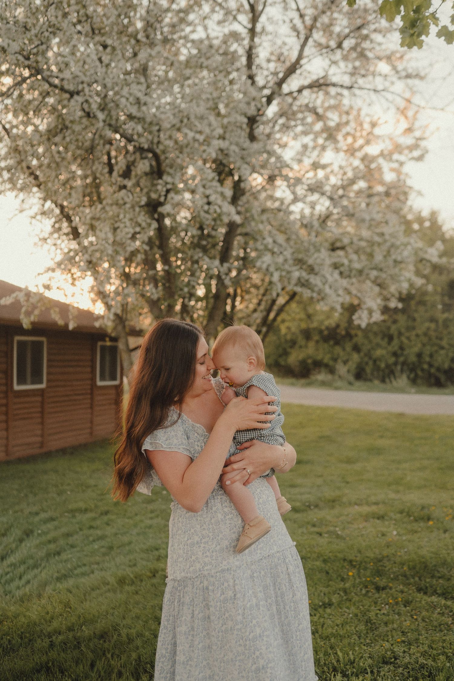 Tender moment of mother in floral maxi dress kissing baby near blooming white tree during warm golden sunset.