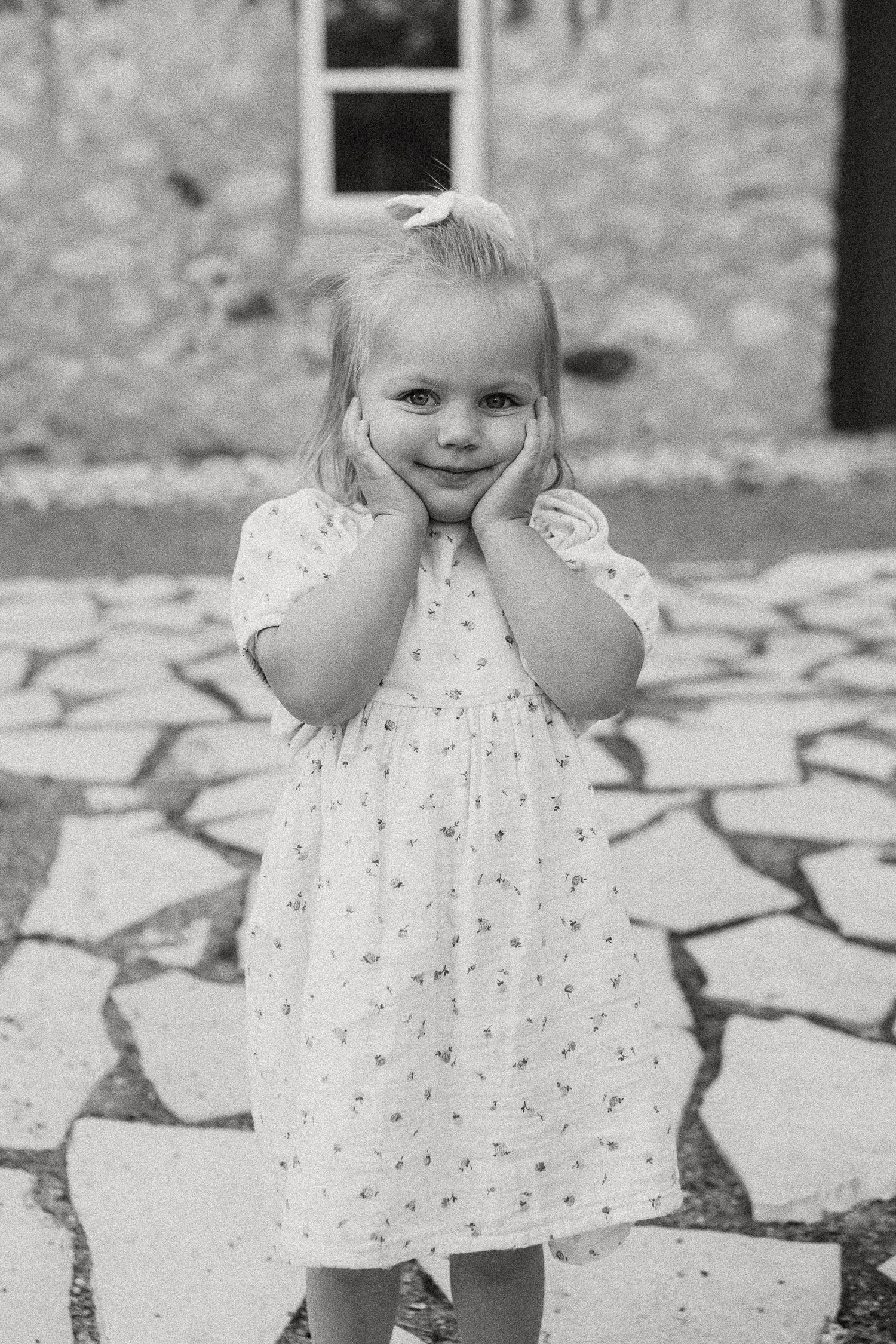 Adorable blonde toddler girl in white floral dress posing with hands on cheeks in black and white photo.