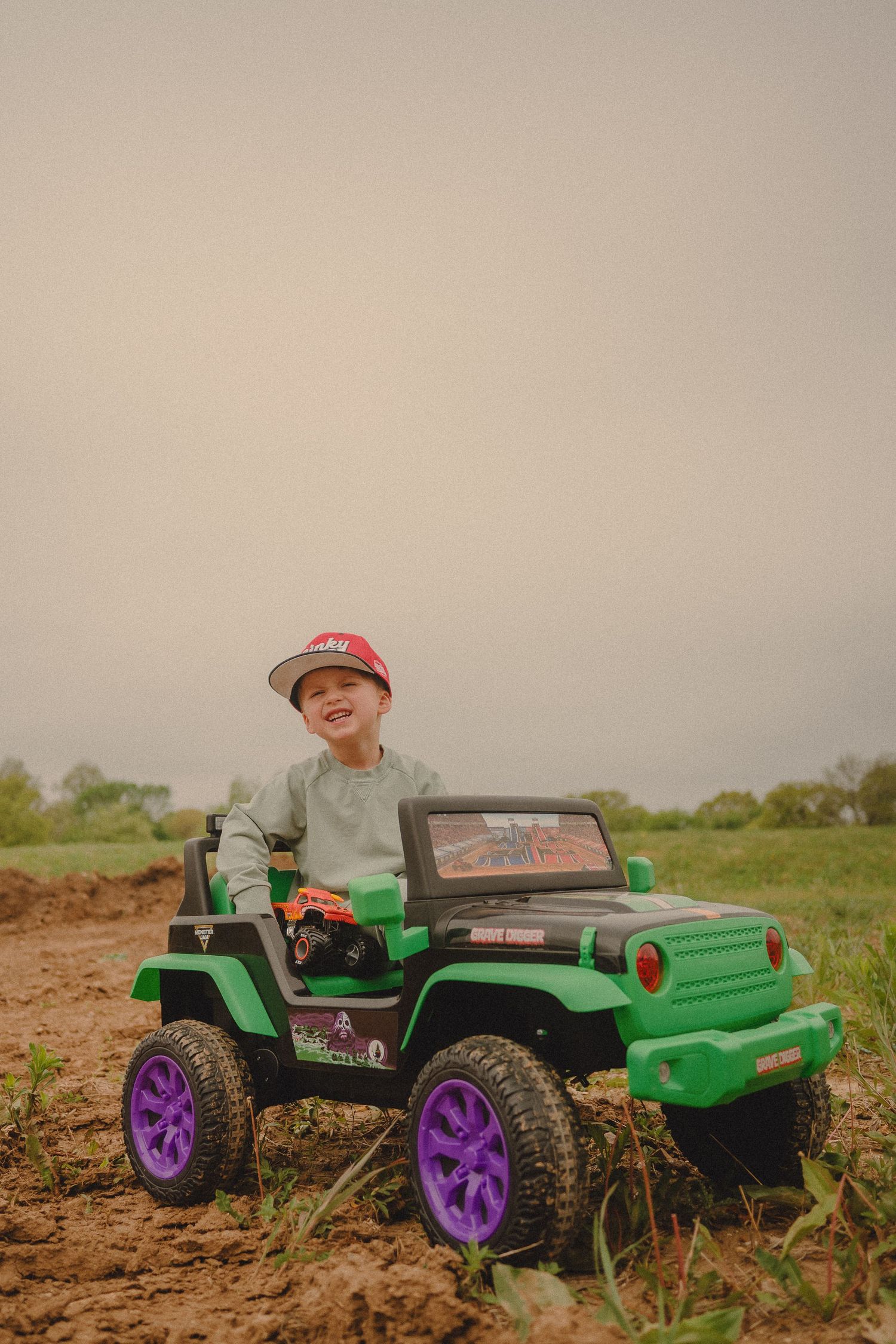 Smiling boy in red cap riding green and purple toy Jeep on dirt path outdoors.