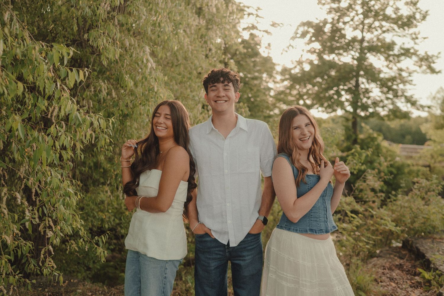 Three smiling young adults posing outdoors among lush green trees in warm golden hour light.