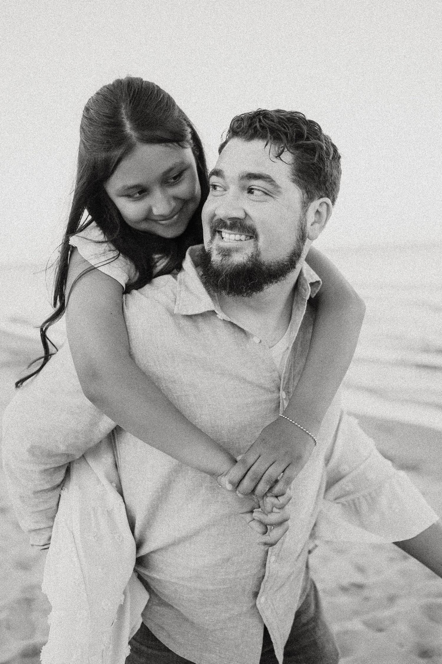 Black and white photo of smiling man giving woman a piggyback ride on a sandy beach.
