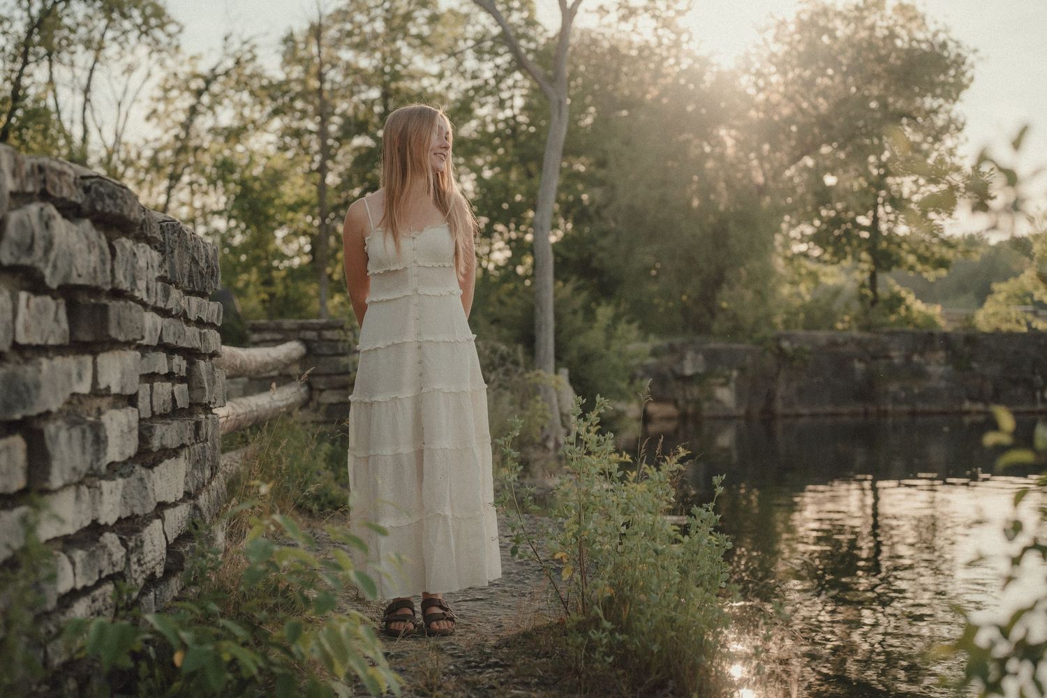 A woman in a white lace maxi dress stands by a stone wall near a sunlit pond surrounded by lush greenery.