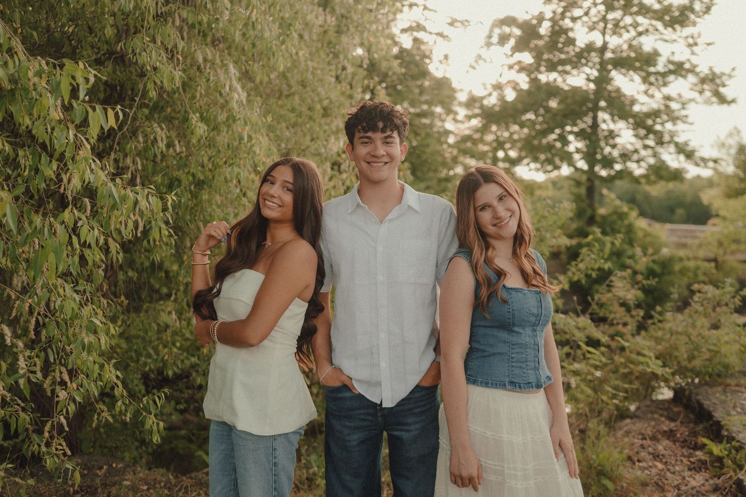 Three young adults smiling together outdoors surrounded by lush green trees in warm golden hour light.