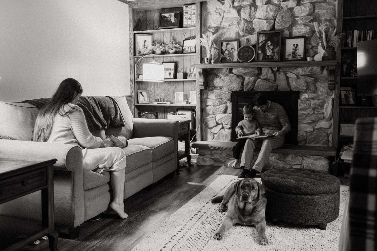 A black and white photo of someone relaxing on a couch near a stone fireplace with a dog resting on the floor.