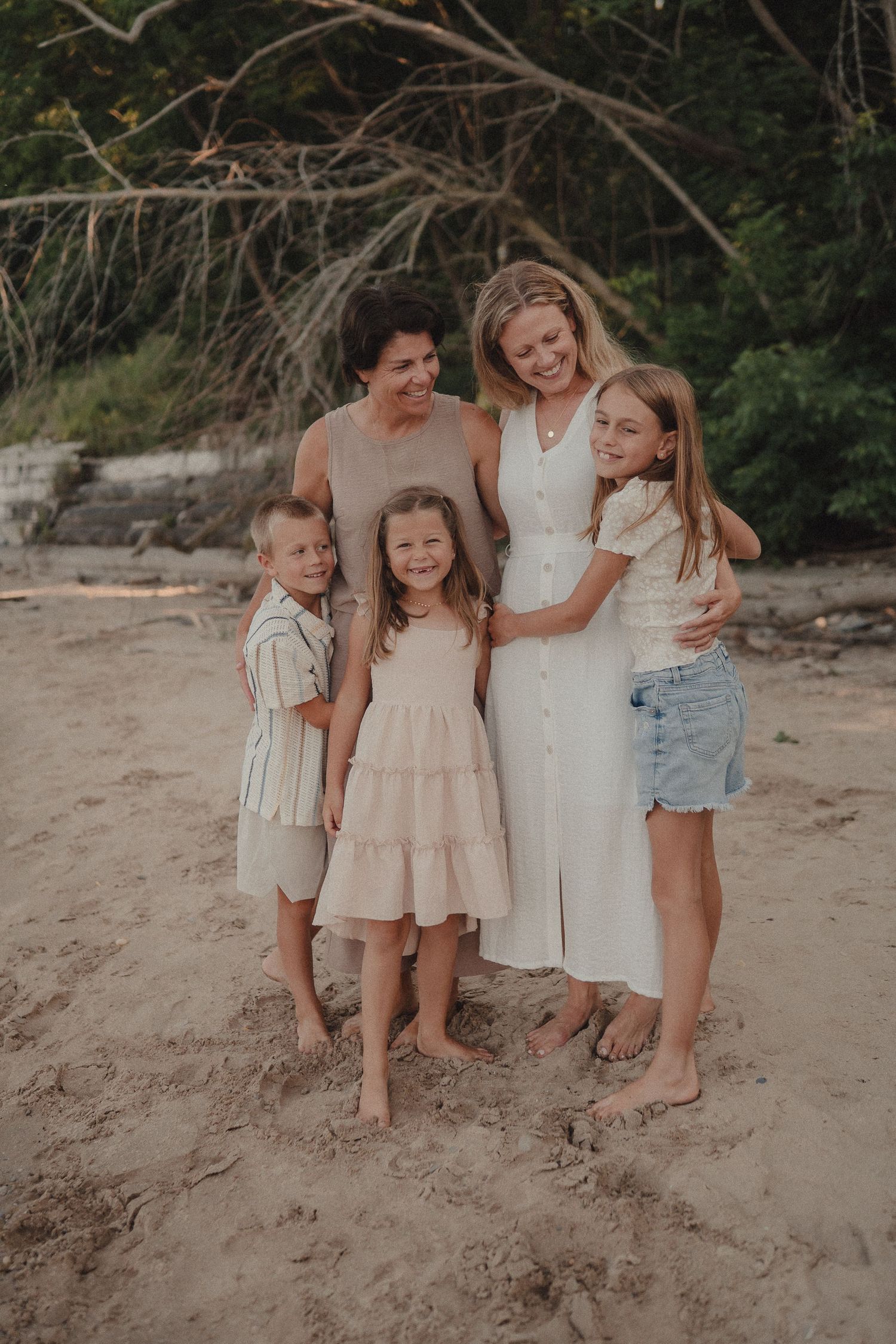 Two women and three children pose together smiling on a sandy beach at sunset.