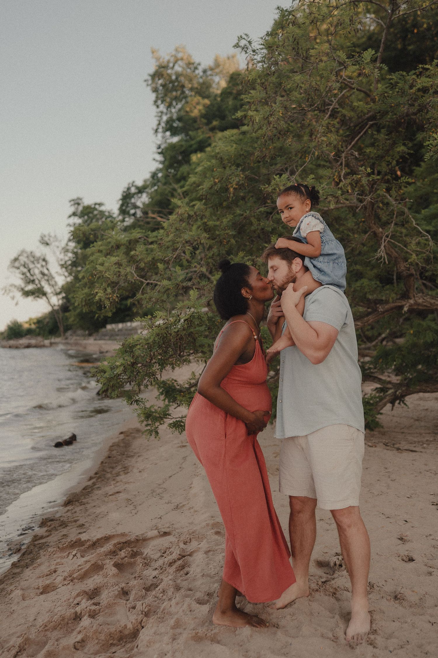 Interracial family on beach, pregnant woman in coral dress kissing man with toddler on shoulders.