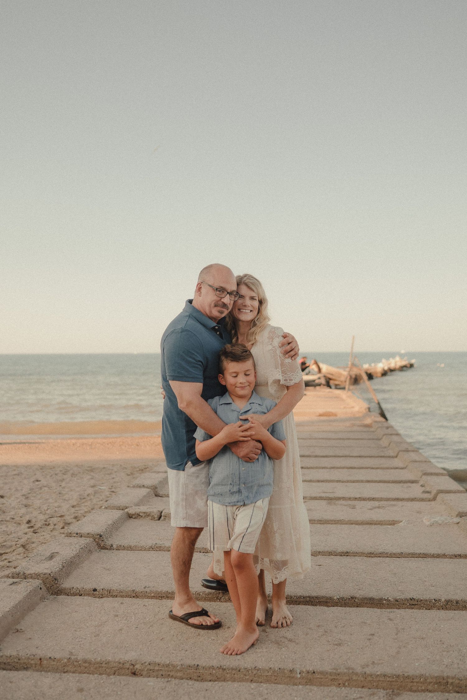 A happy family of three poses together on a beach pier, embracing warmly with the ocean behind them.