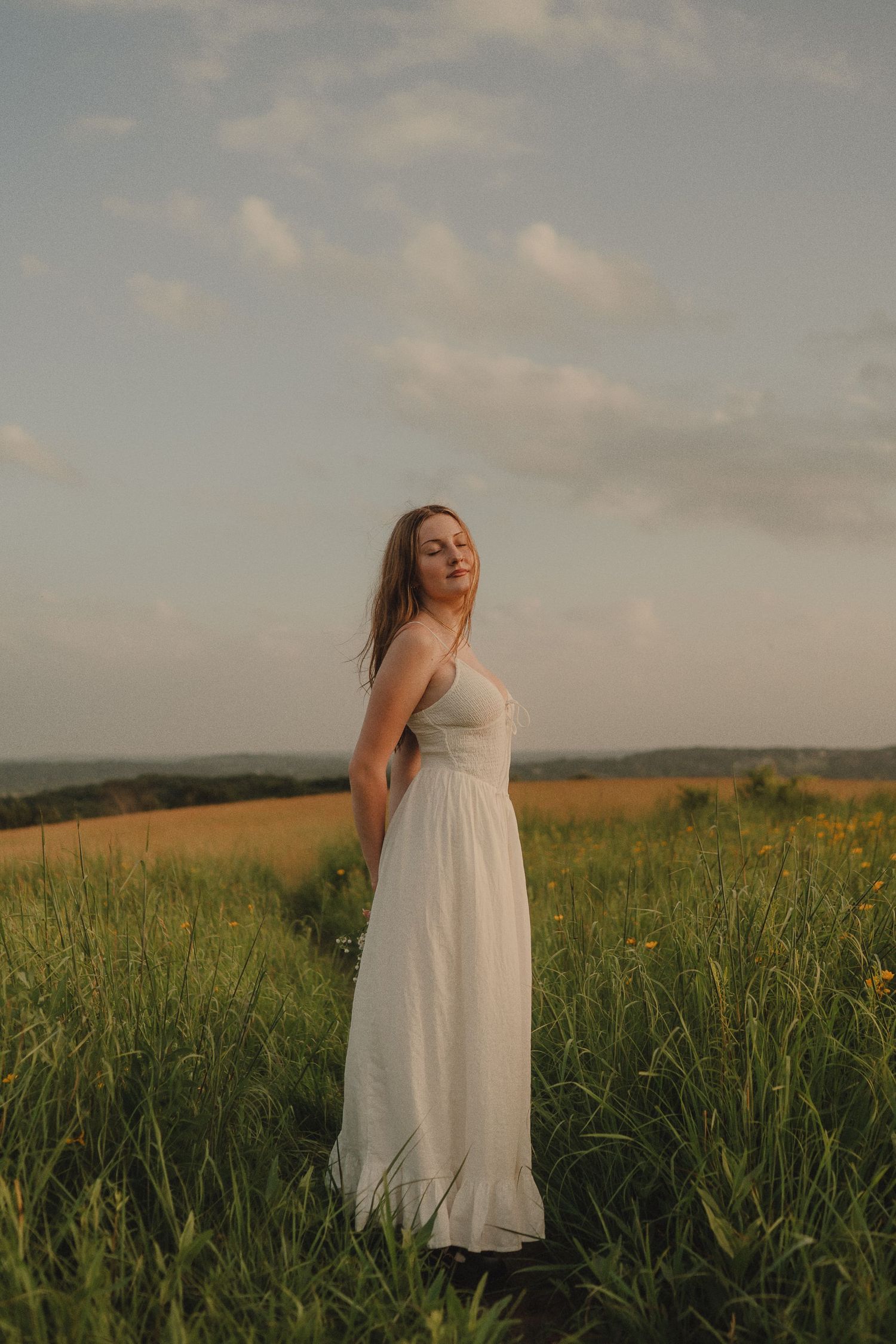 Young woman in long white dress standing in green meadow at golden hour with cloudy sky backdrop.
