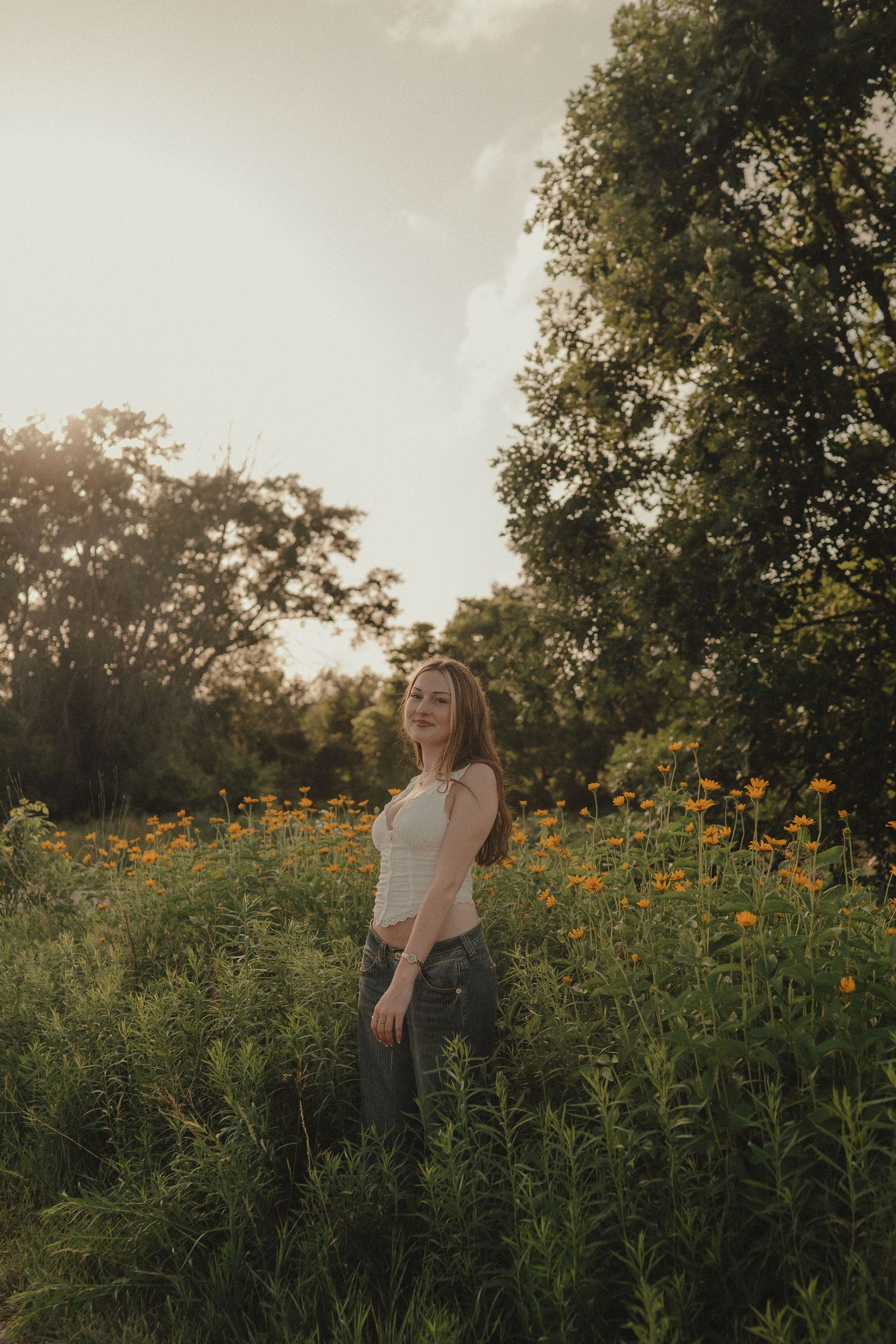Young woman in white crop top and jeans standing in a field of orange wildflowers at golden hour.