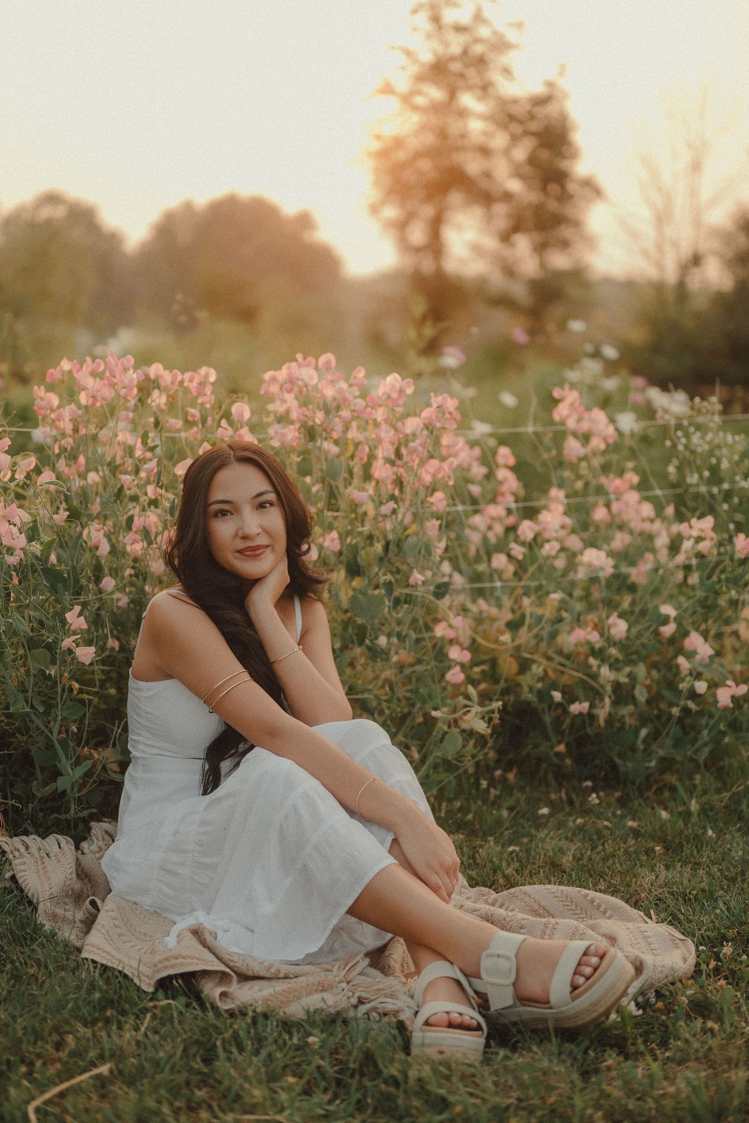 Young woman in white dress sitting relaxed among pink wildflowers in a meadow at sunset.