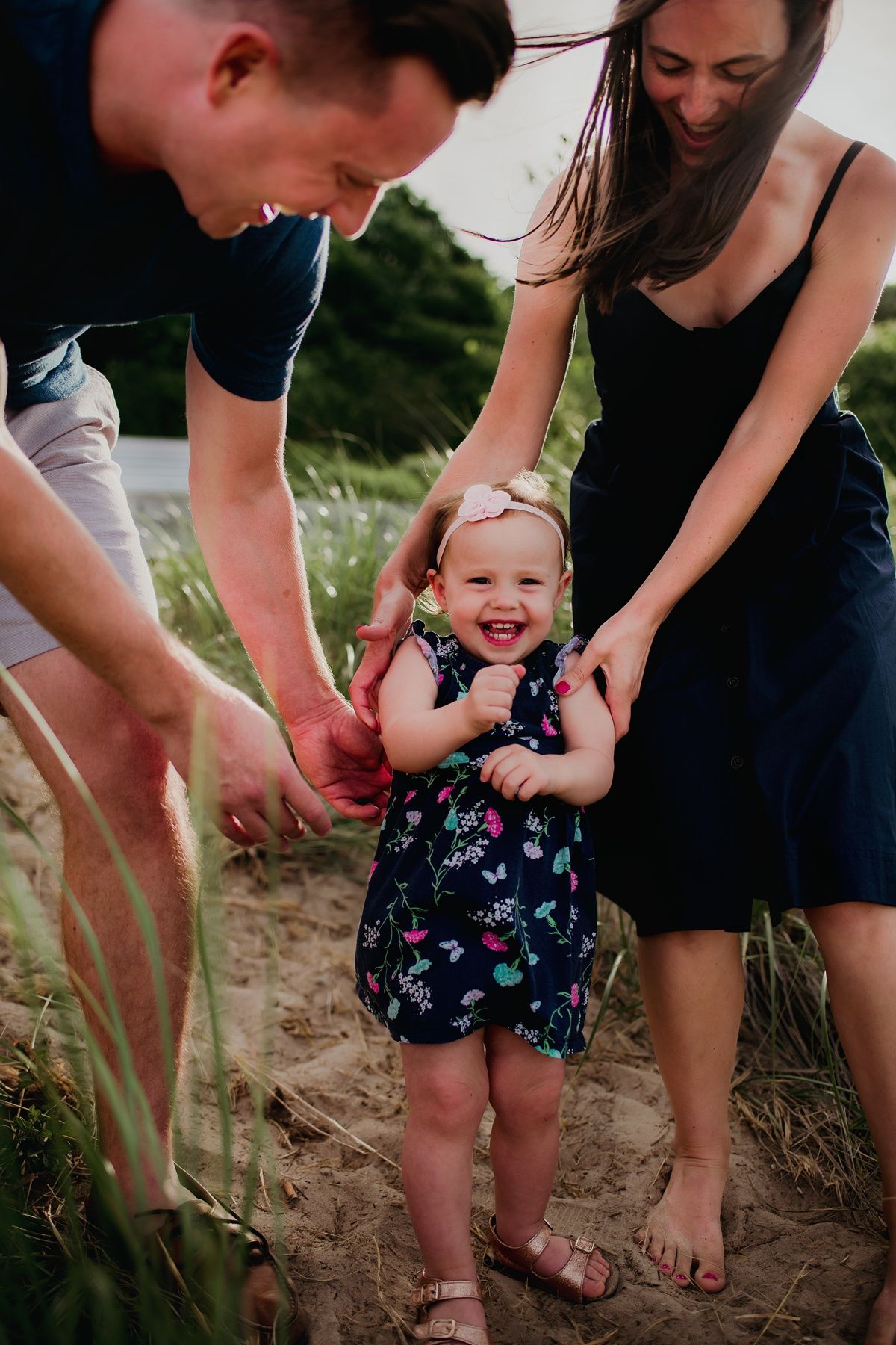 A happy toddler in a floral dress laughs while playing with parents on a sandy path.