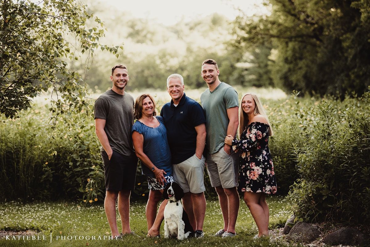 An outdoor family portrait with a black and white dog in a lush green setting.