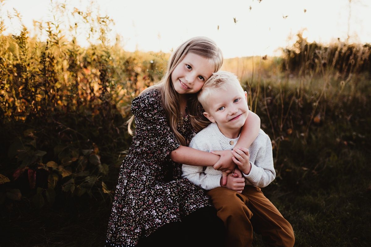 Siblings share loving embrace while posing in golden evening light among tall grass.