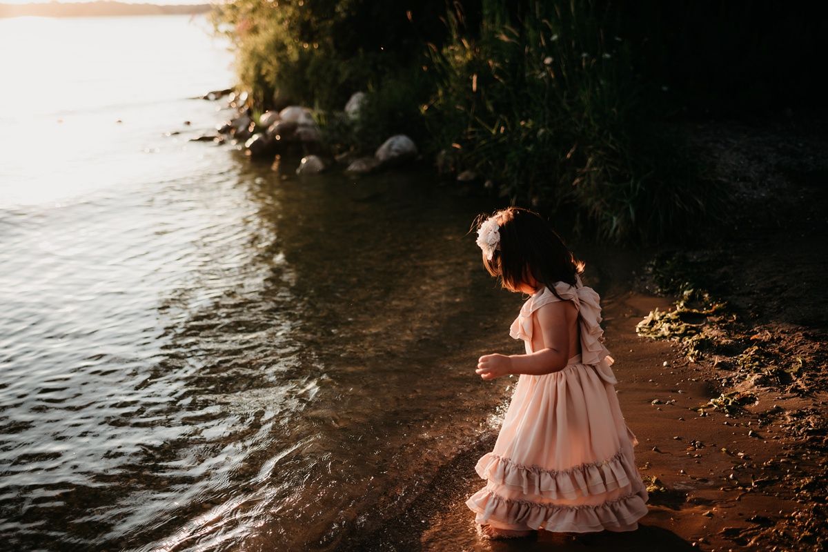 Child in ruffled pink dress explores rocky shoreline at golden hour.