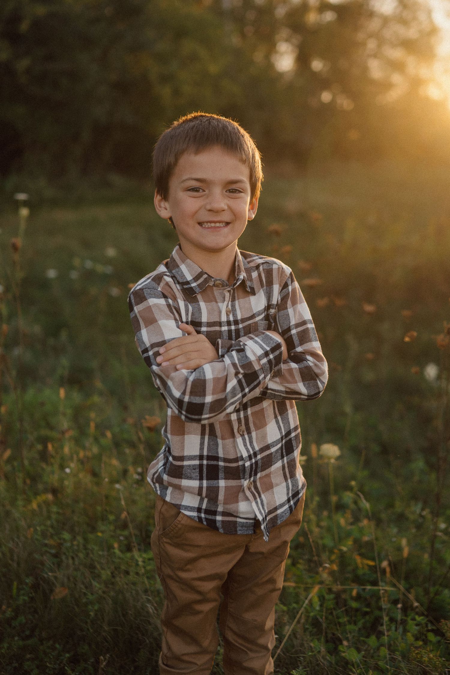 A plaid shirt and brown pants are worn in this golden hour outdoor portrait against a natural background.