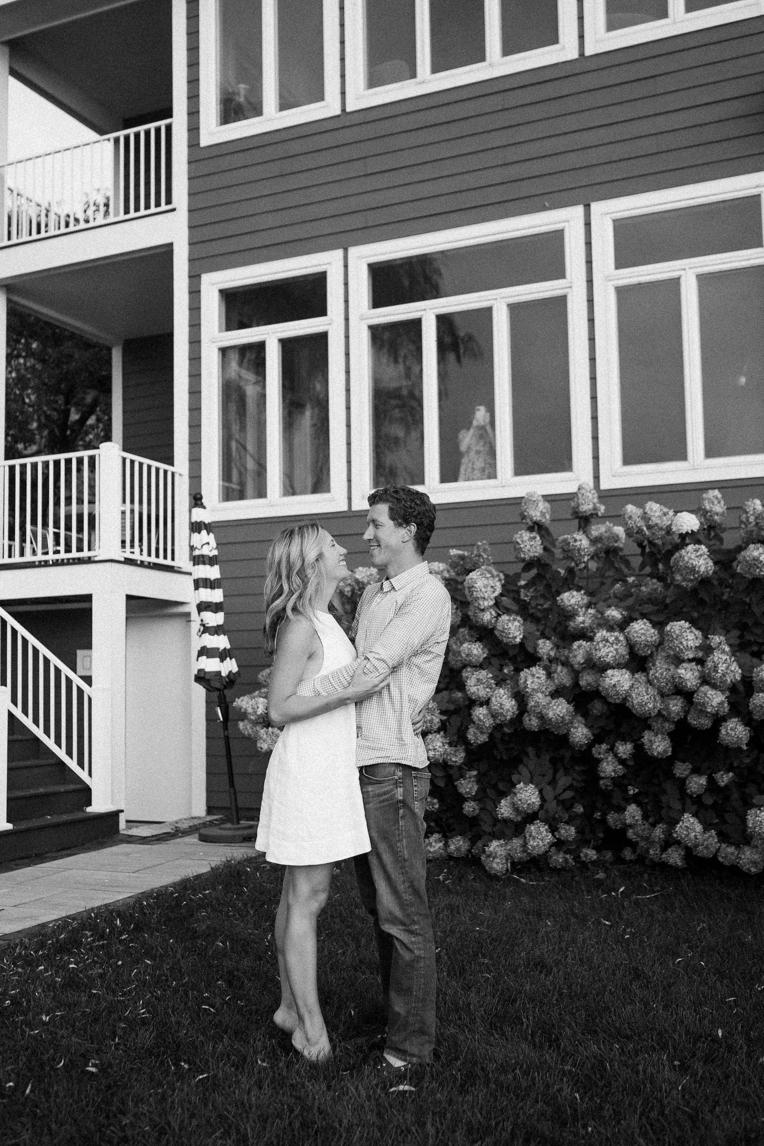 A couple dances playfully on the lawn in front of a modern house with hydrangea bushes.