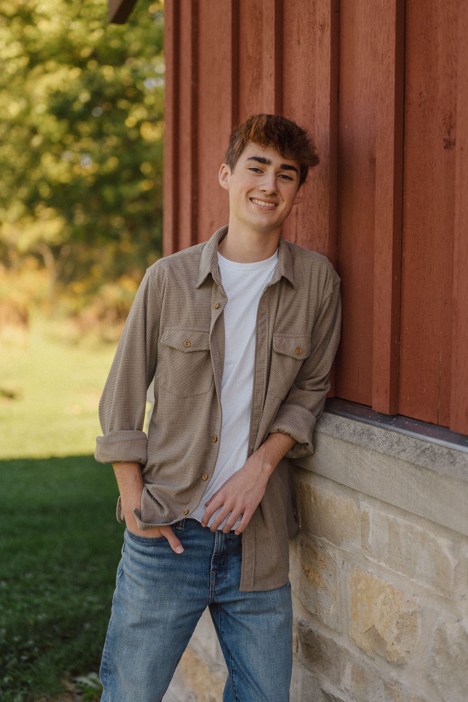 A casual outdoor portrait against a red barn wall and stone foundation in warm natural lighting.