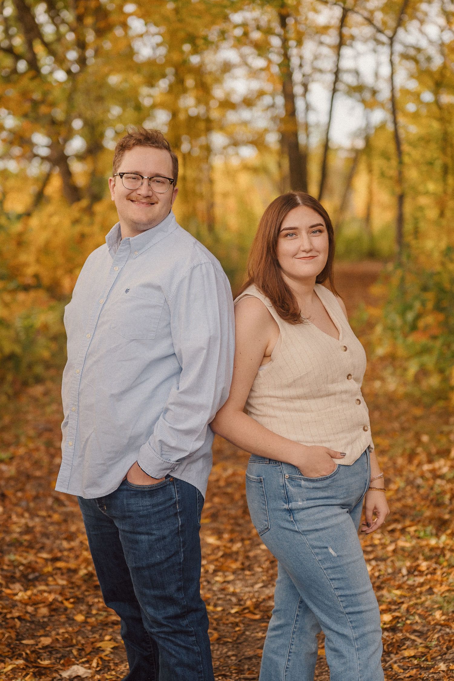 A couple poses playfully in autumn woods with golden leaves and sunlight filtering through the trees.