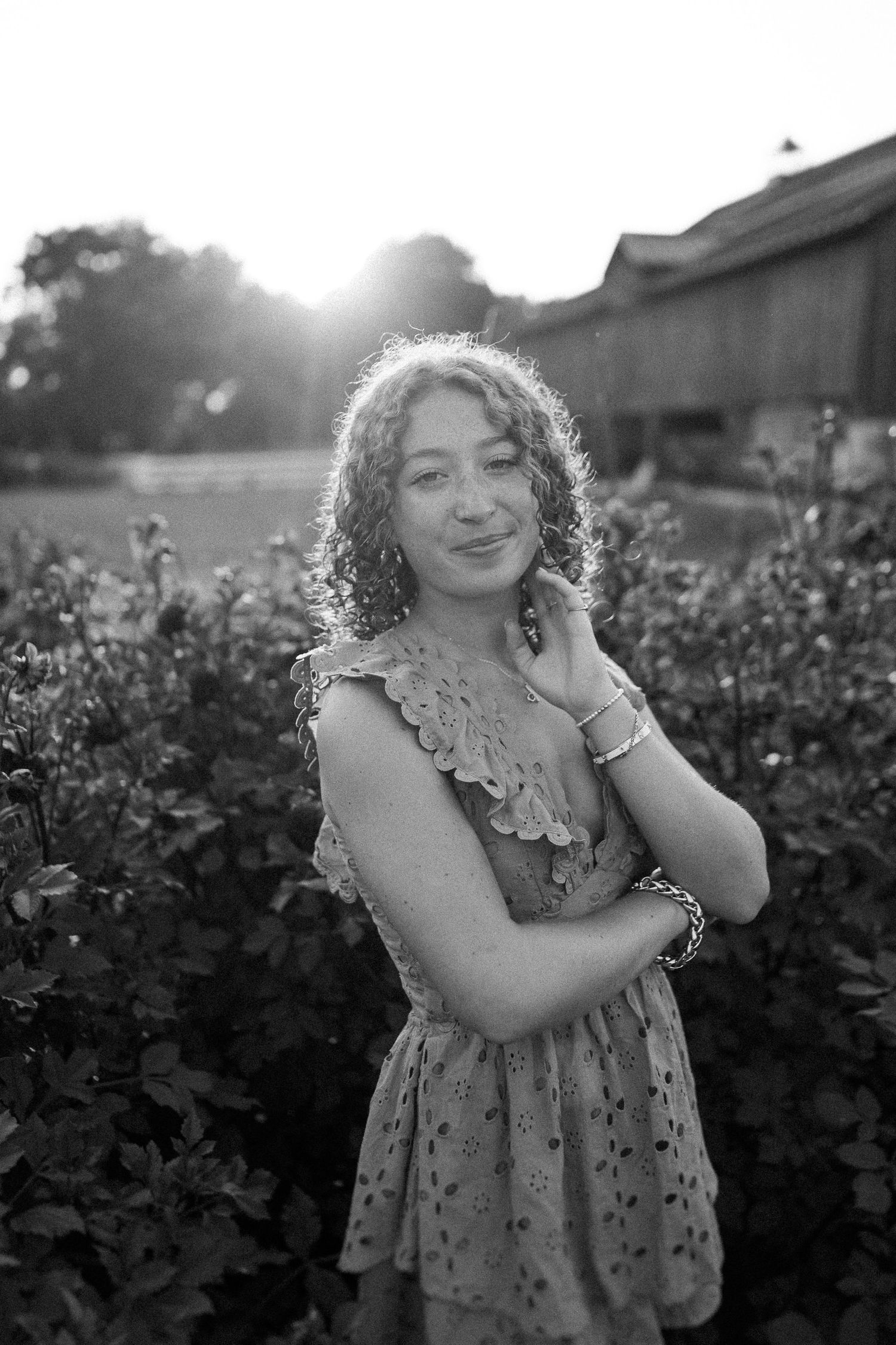 Black and white portrait of a person in a floral dress standing in a garden at sunset with backlit hair.