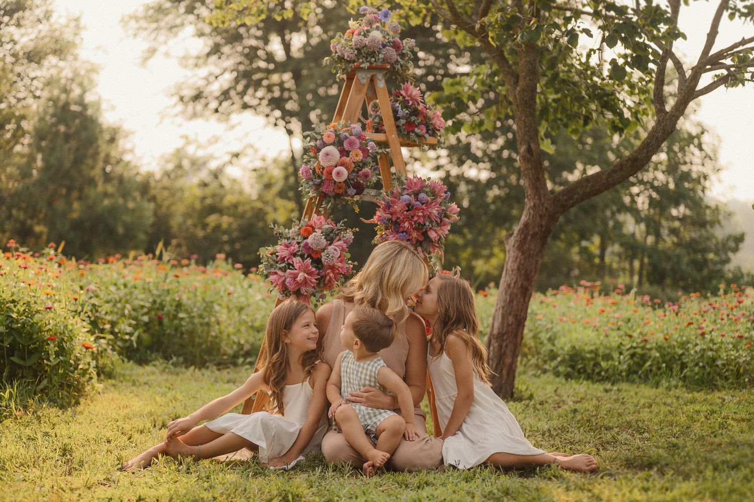People sitting together on grass near a wooden ladder decorated with pink flowers in a sunny garden setting.