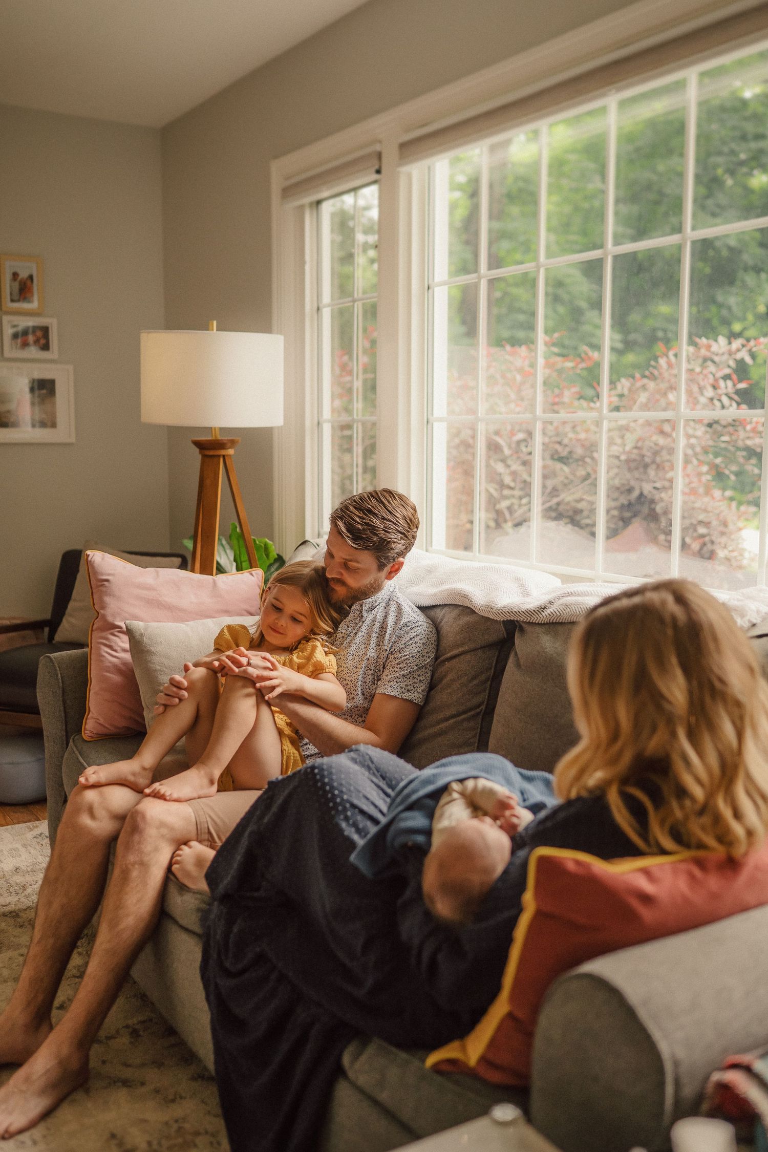 A relaxed family lounging together on a couch in a sunlit living room with large windows.