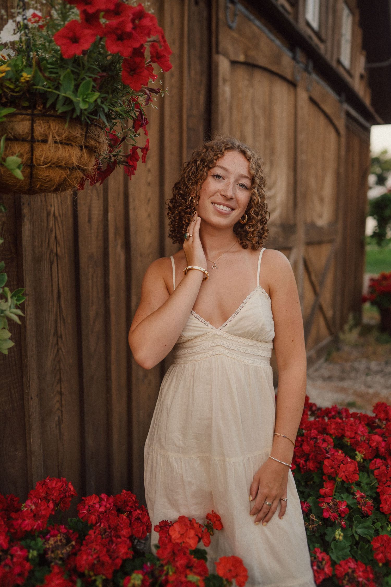 A woman in a white sundress poses by a rustic barn wall with vibrant red flowers and hanging baskets.