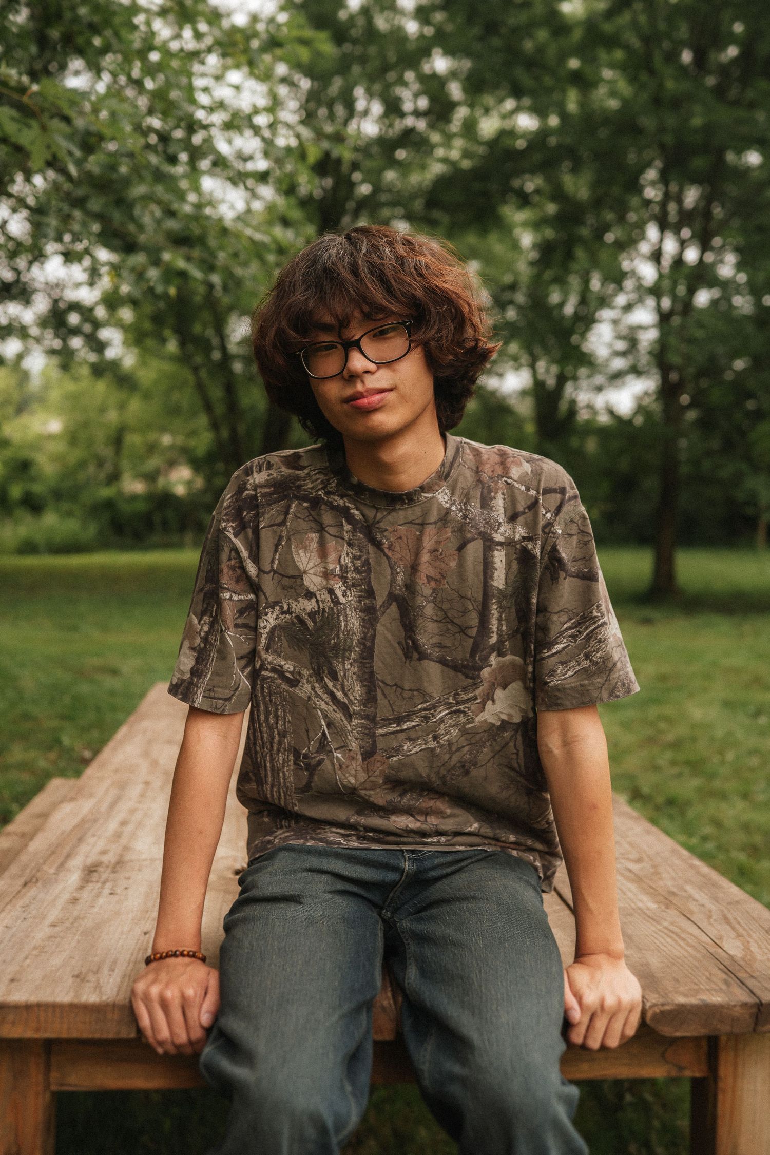 A person wearing a camouflage t-shirt and glasses sits on a wooden bench in a lush green outdoor setting.