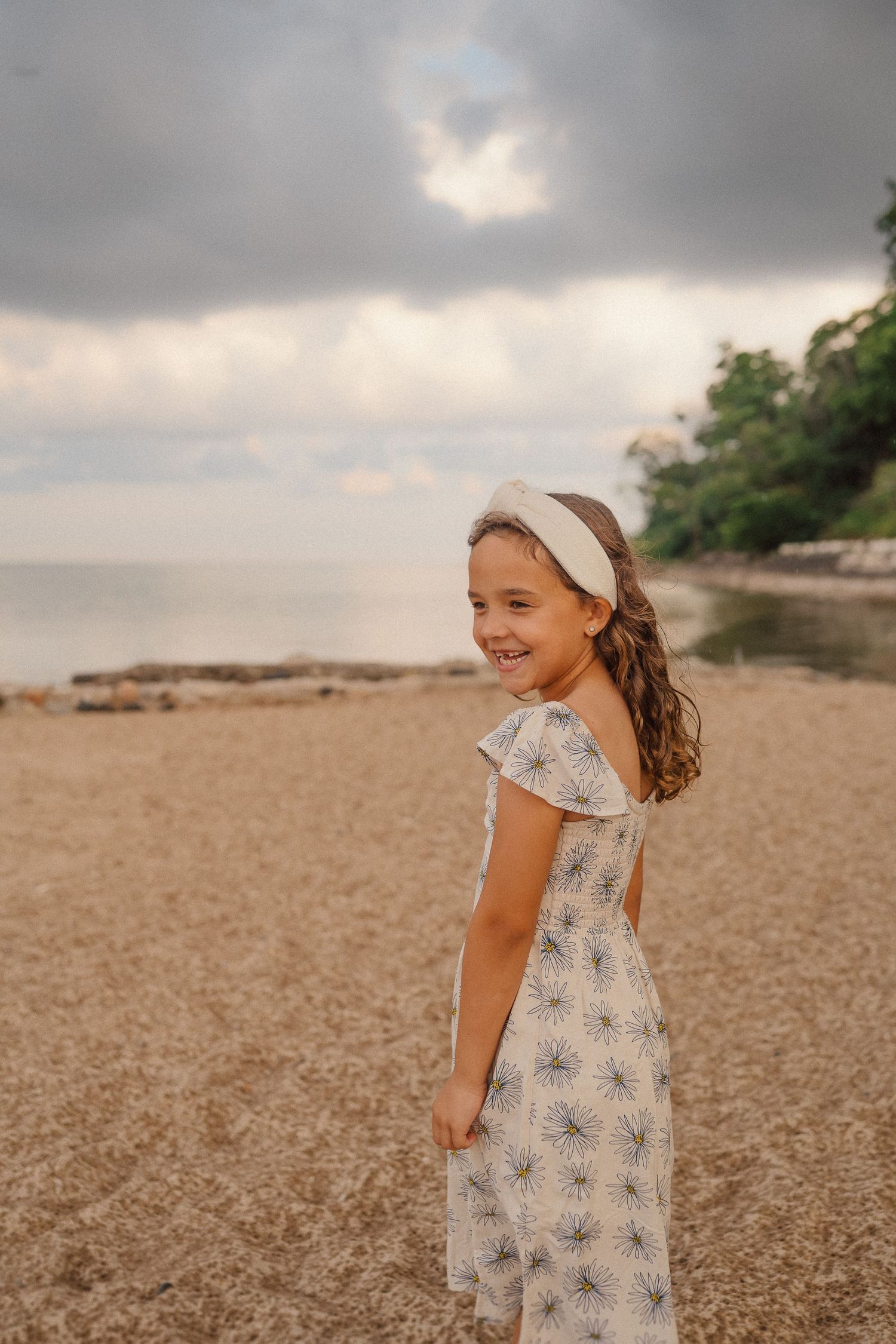 A young girl in a white floral dress smiles on a sandy beach with stormy skies above.