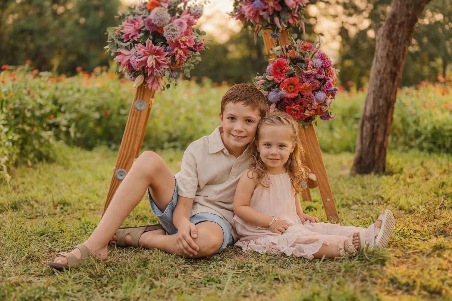 Children sitting under wooden ladder arch adorned with pink and red floral arrangements in garden setting.