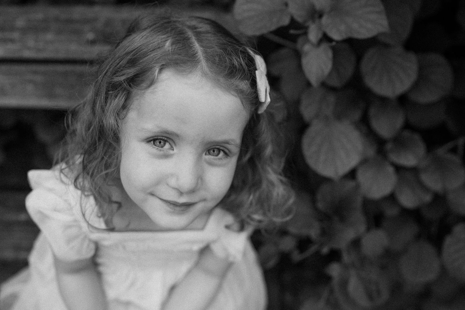 Black and white portrait of a young child wearing a light-colored dress with leaves visible in the background.