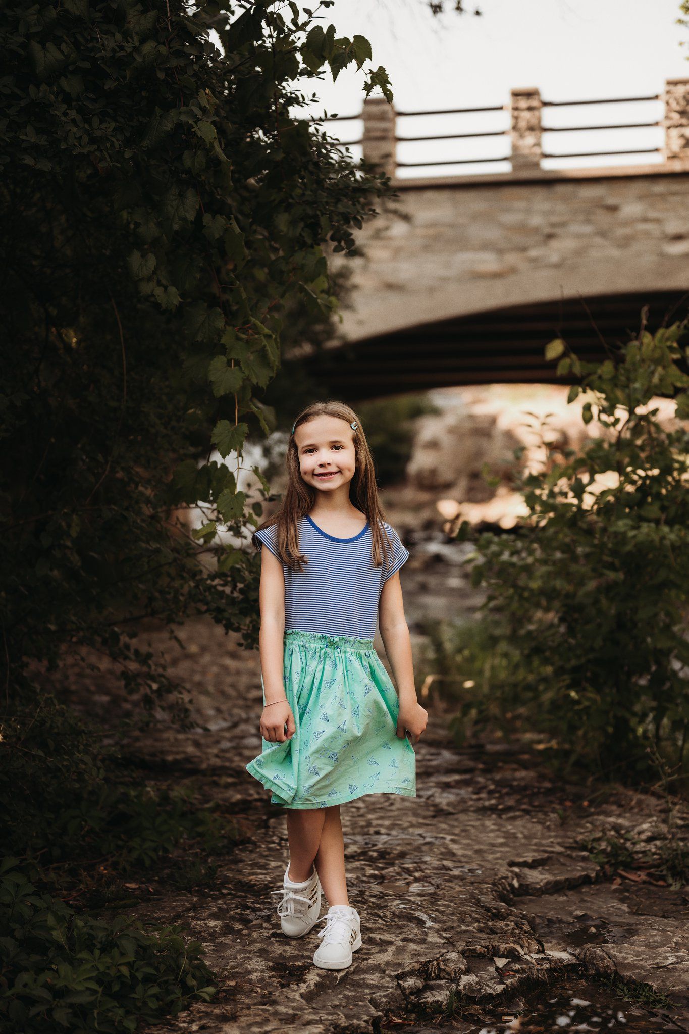 Child in striped top and mint green skirt standing on path under stone bridge.