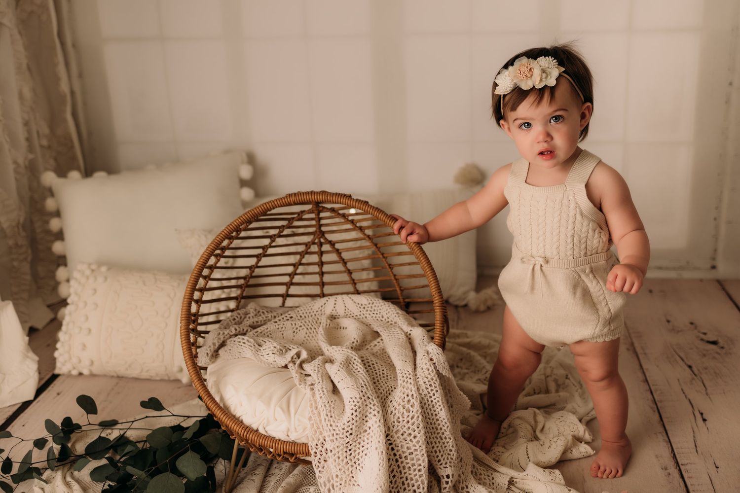Baby stands next to rattan chair holding edge while wearing cream outfit with floral headband.