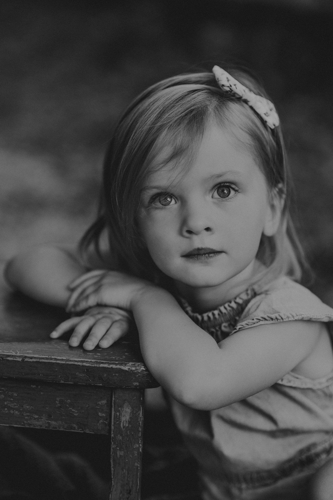 Black and white portrait of child leaning on wooden furniture with a hair bow.