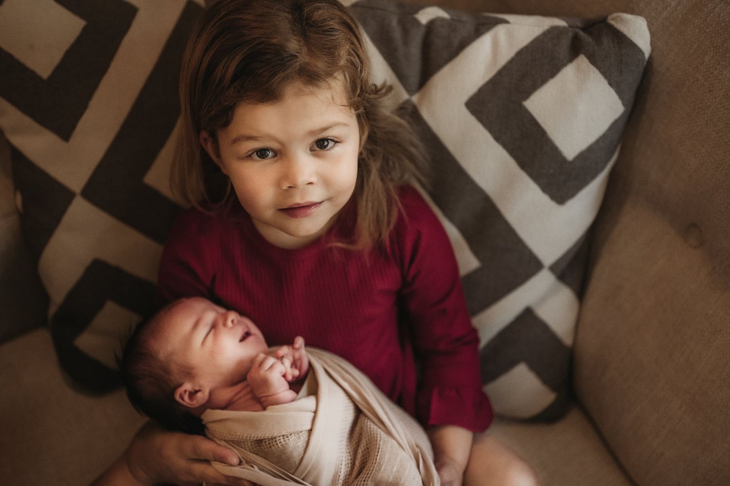 Girl in burgundy sweater sits with newborn sibling on couch with patterned pillows.