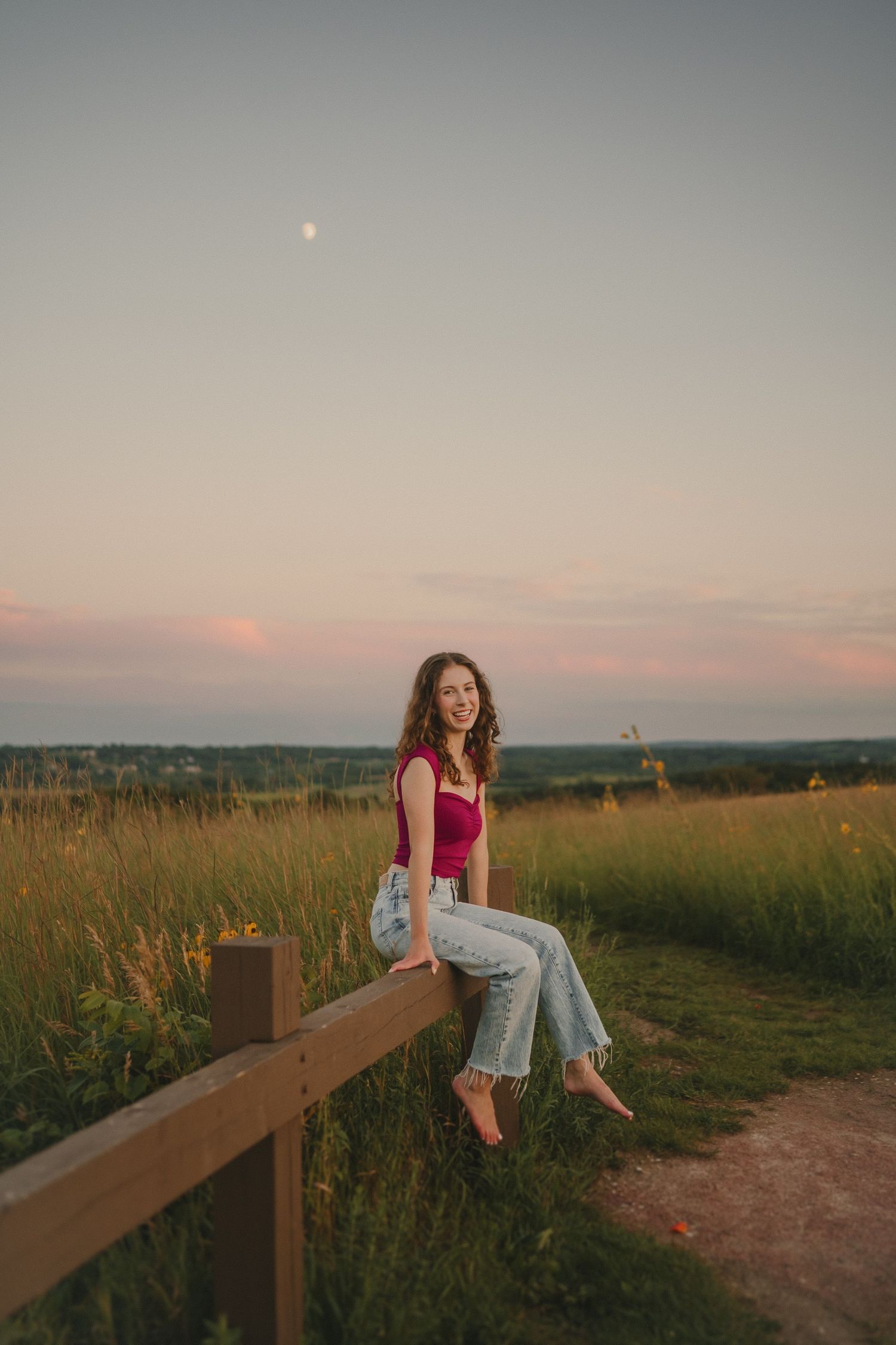 A sunset portrait captures someone sitting on a wooden fence with rolling hills in the background.
