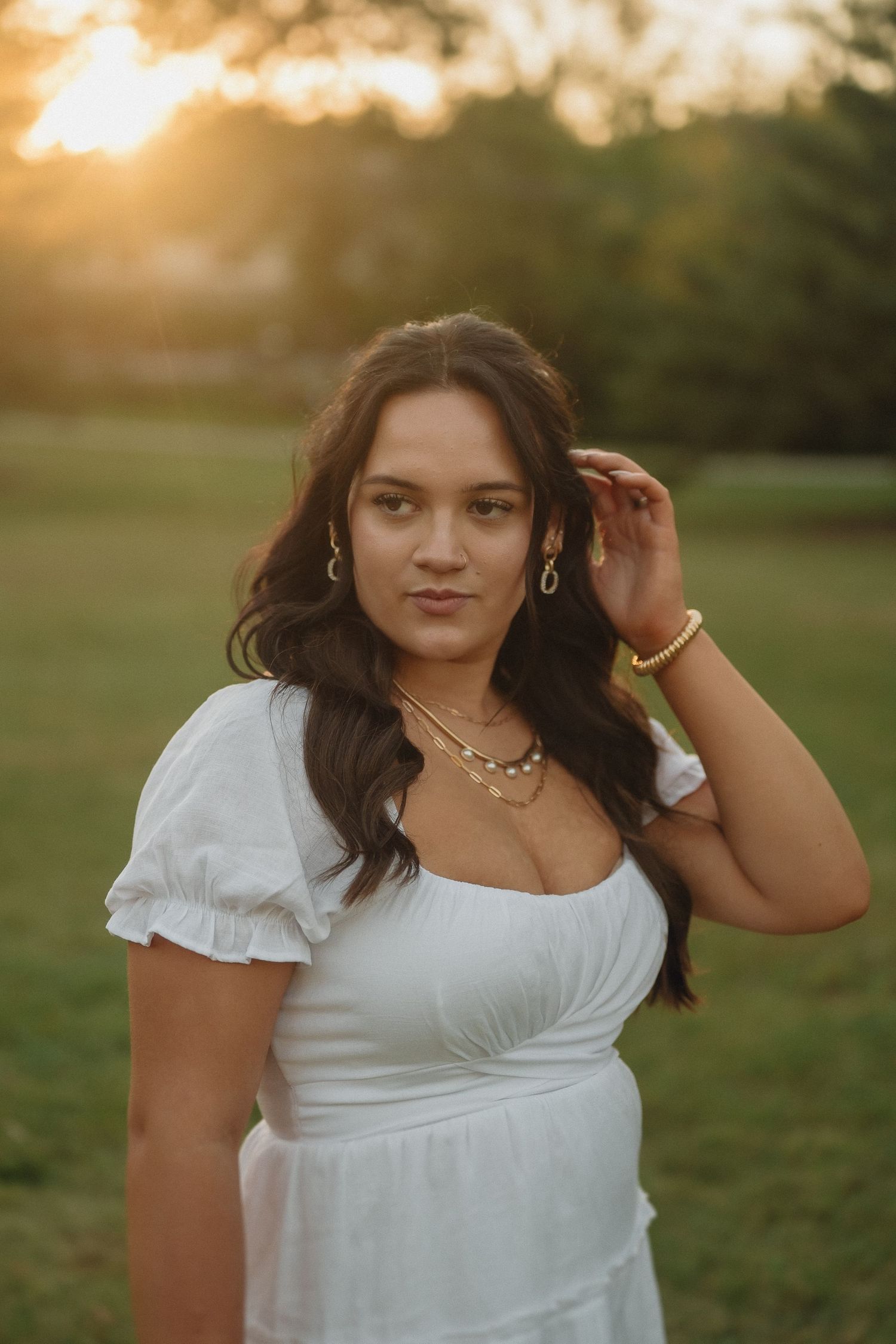 A person in a white dress poses outdoors during golden hour sunset with jewelry and flowing dark hair.