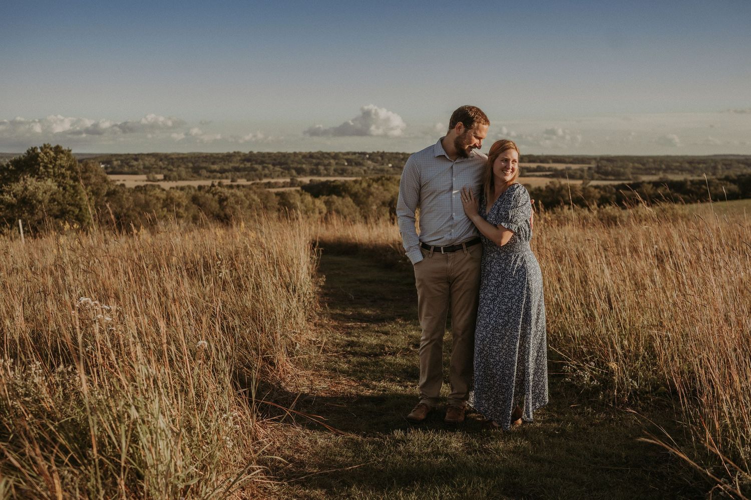 Romantic couple standing together on a dirt path through tall grass at sunset.
