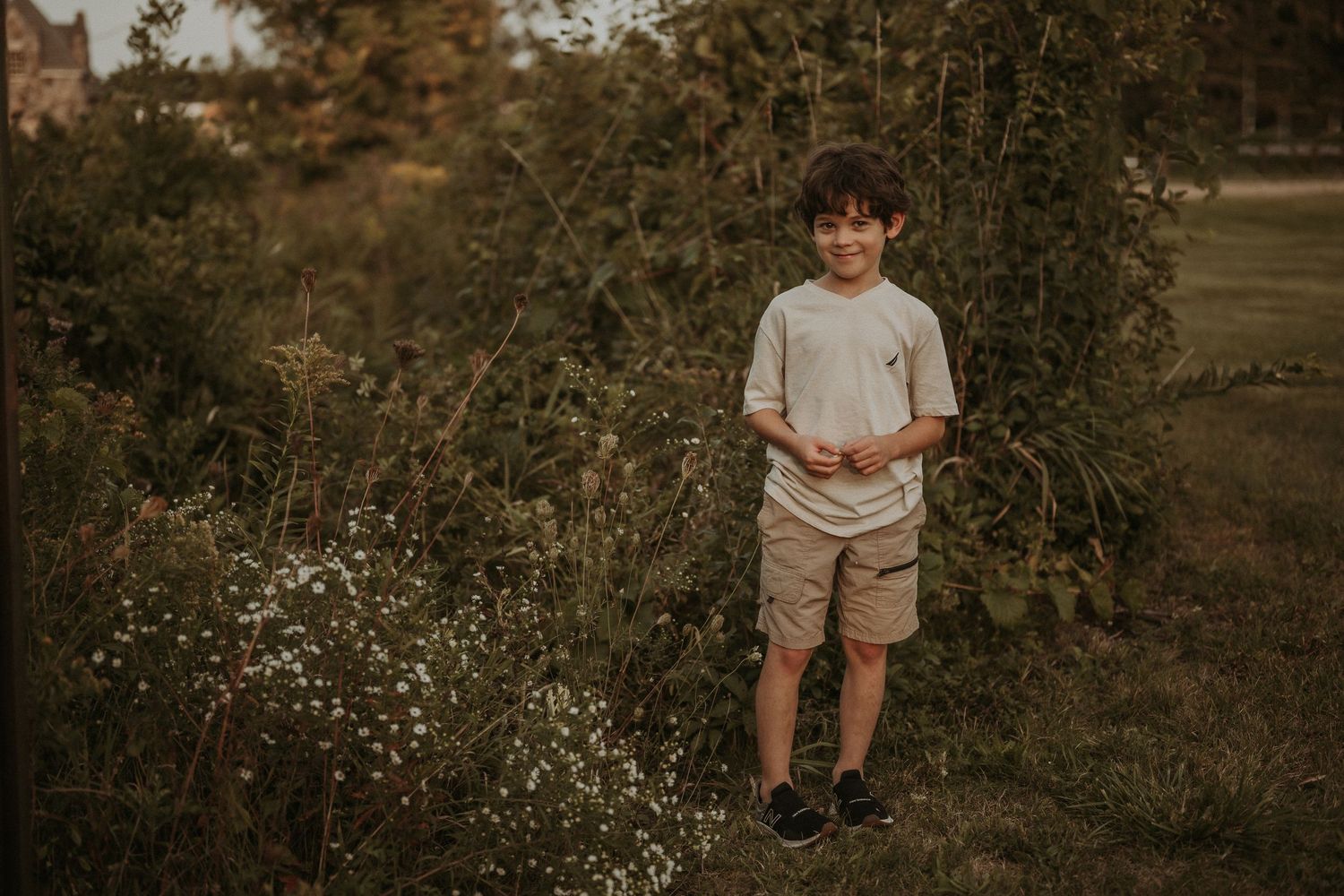 A young boy stands in a natural setting with wildflowers during golden hour light.