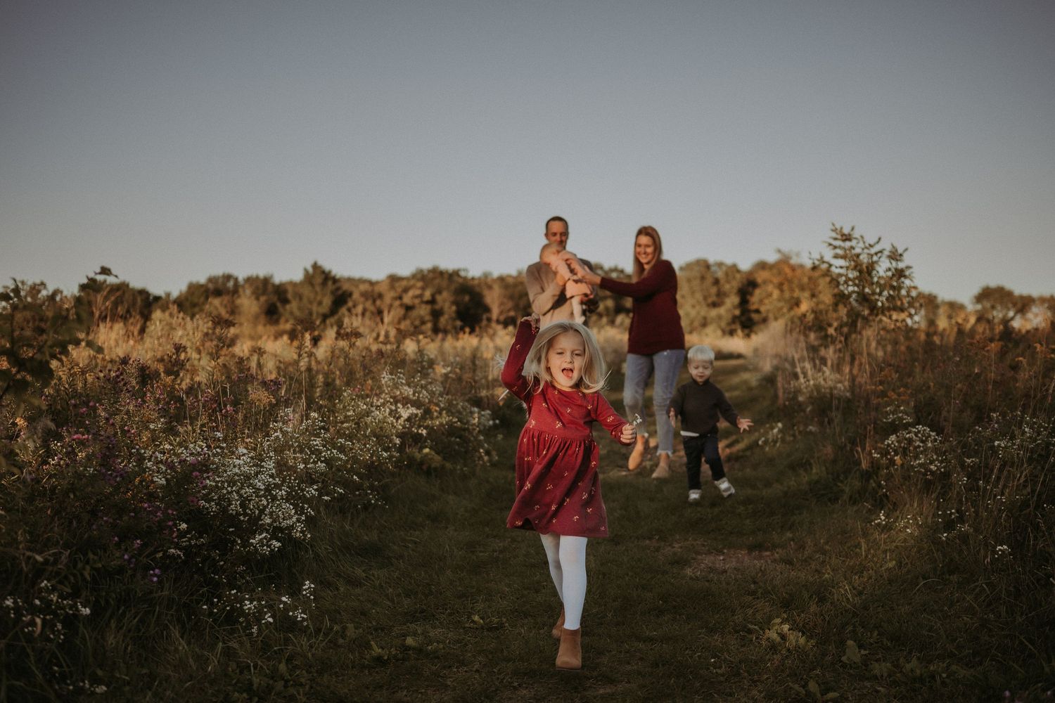 Family walking through wildflower meadow during golden hour with rustic barn in distance.