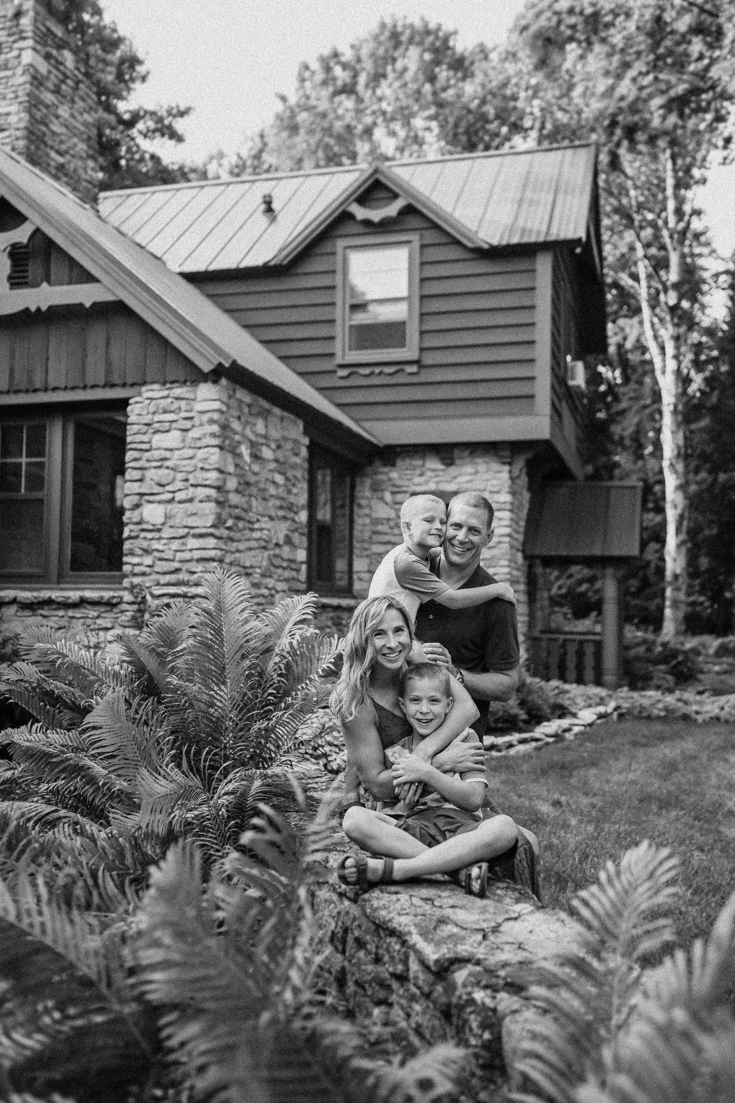 Black and white portrait of family gathered by stone wall with cabin in background.