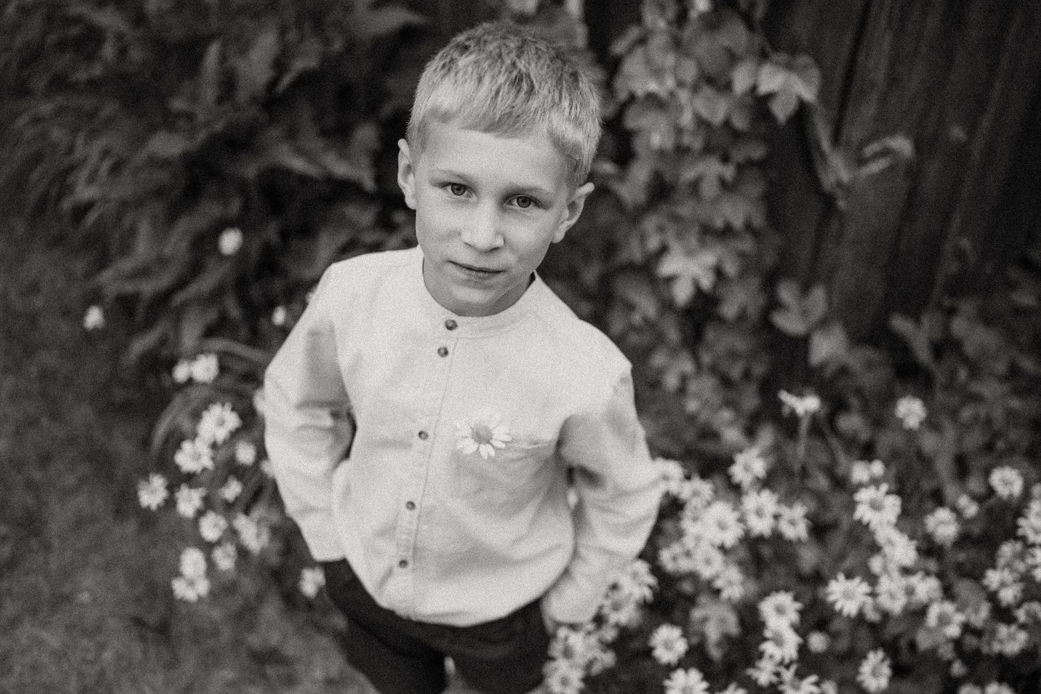 Black and white portrait of a child in a collared shirt standing among garden flowers.