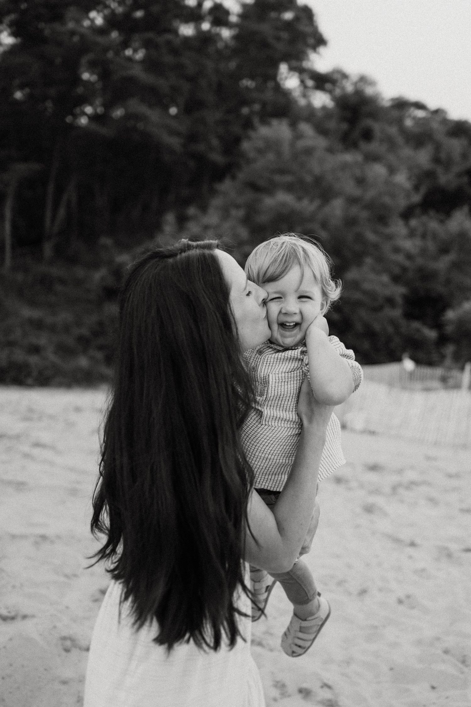 A tender black and white moment between mother and laughing toddler at the beach with trees in background.