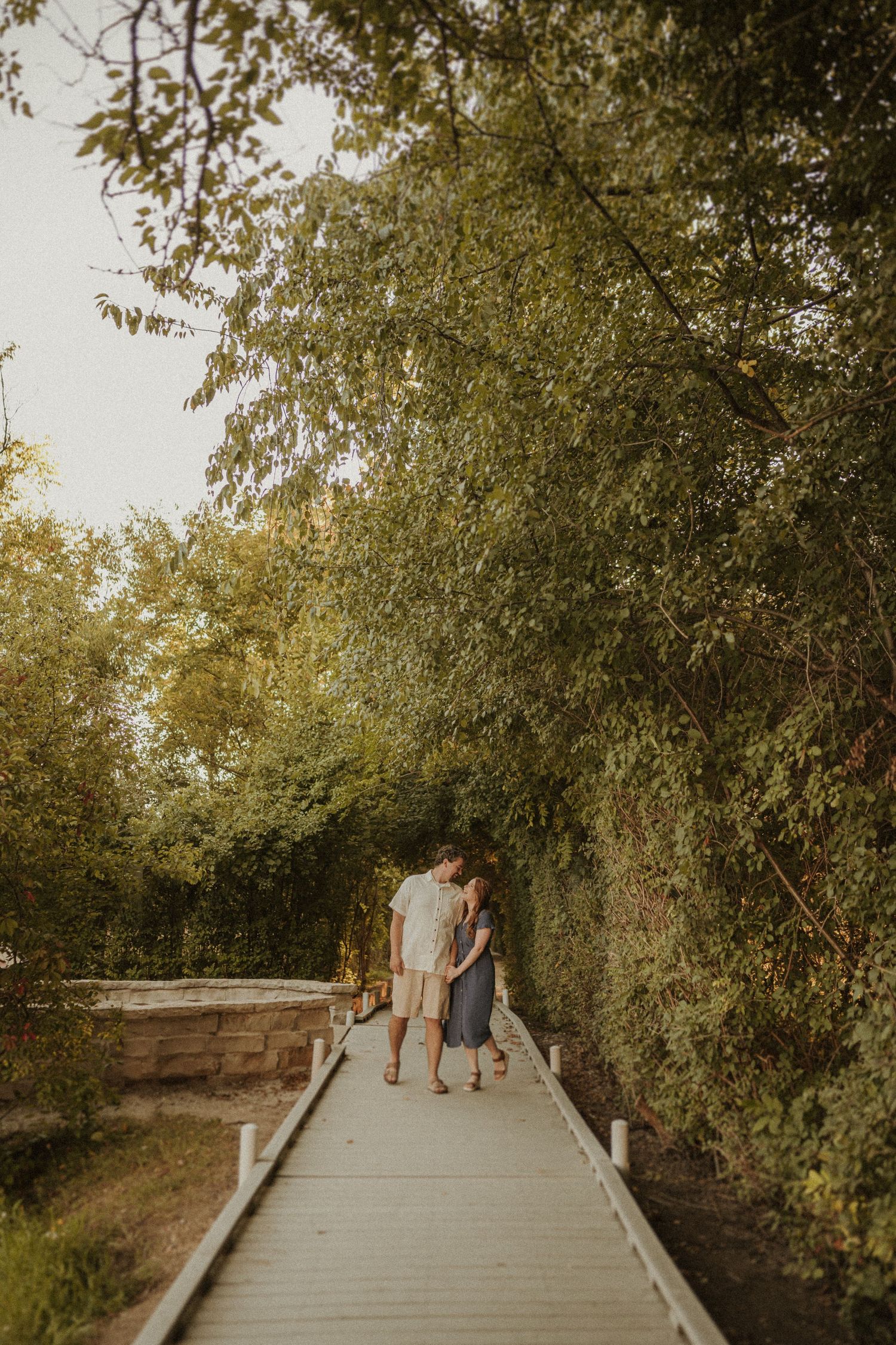 Couple walking hand in hand on a wooden boardwalk surrounded by lush green trees at sunset.