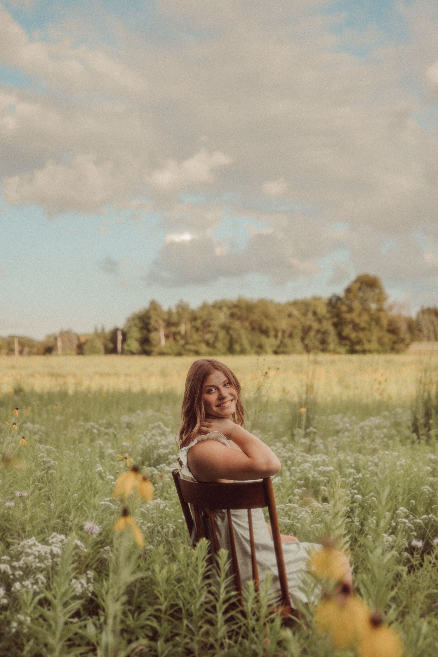 Figure seated in wooden chair among wildflowers during golden hour in rural field.