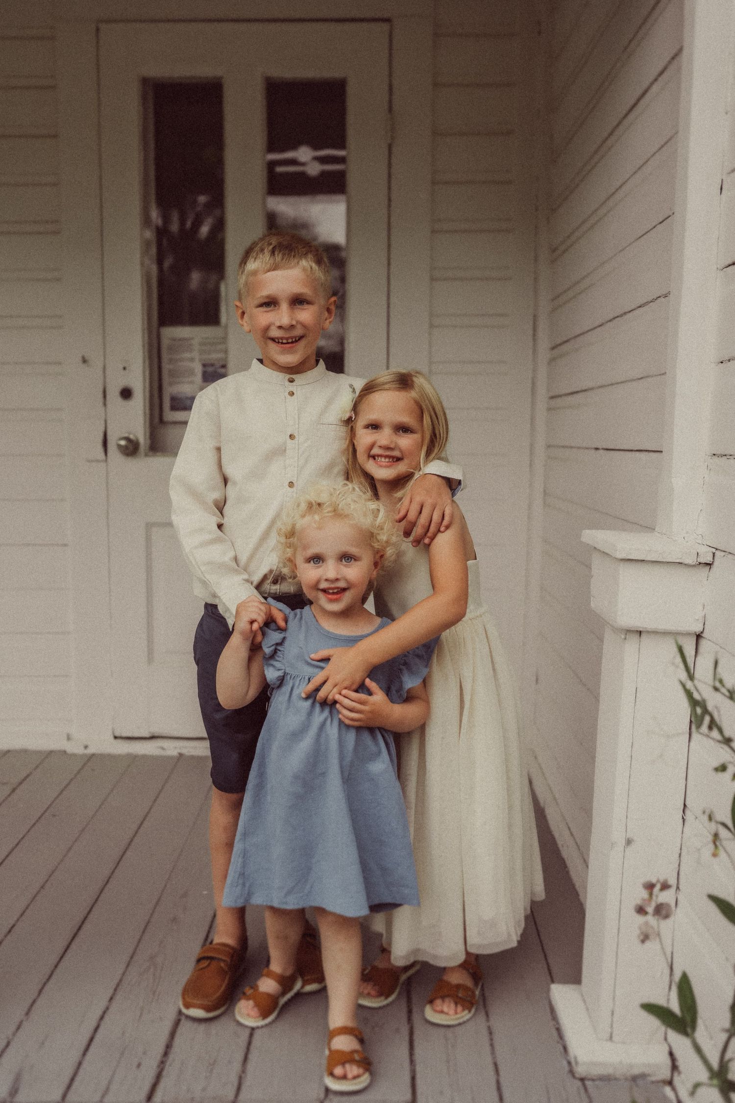 Three siblings pose together on a white farmhouse porch wearing light-colored clothing.