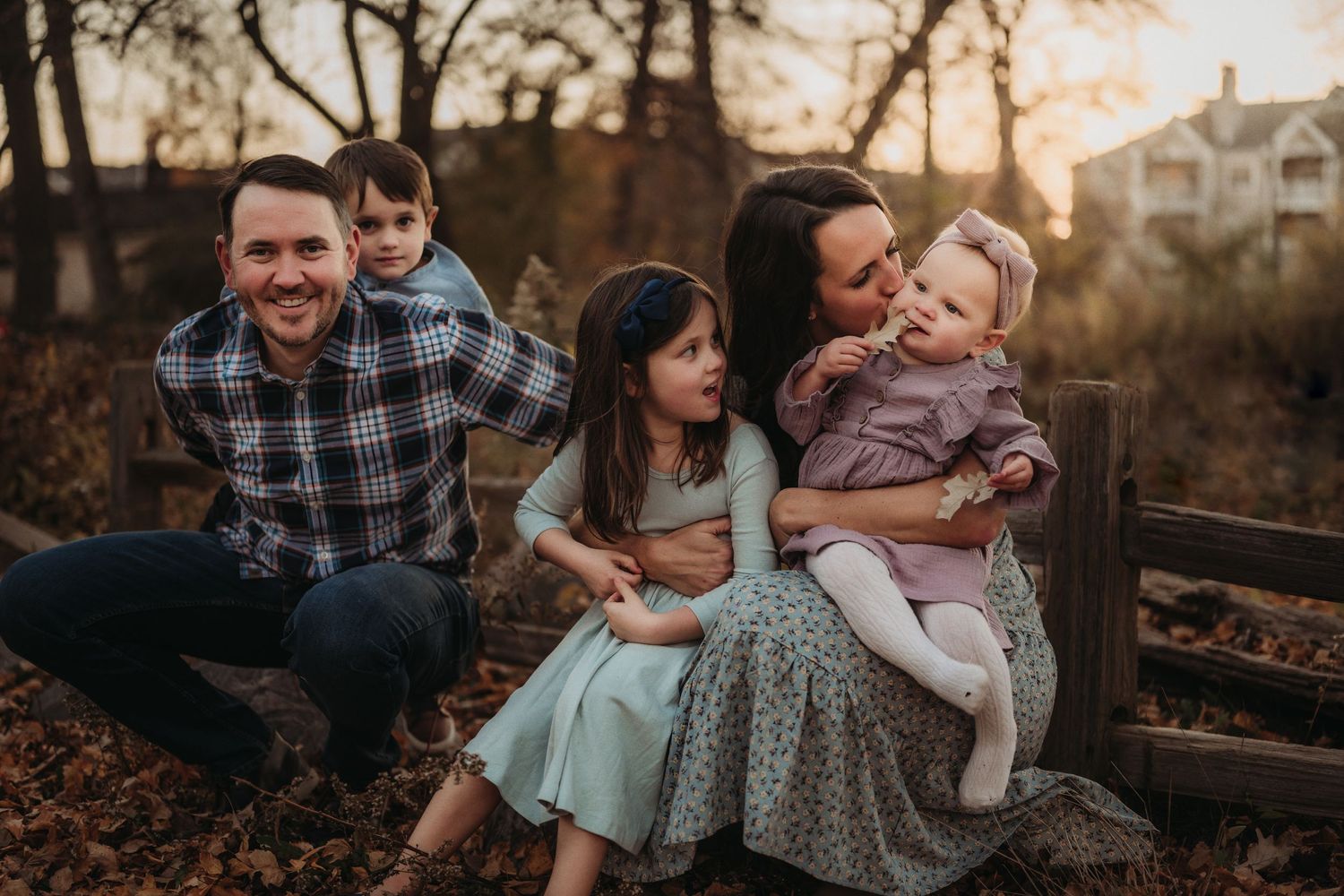 A family shares a loving moment together while sitting on a wooden fence at sunset.