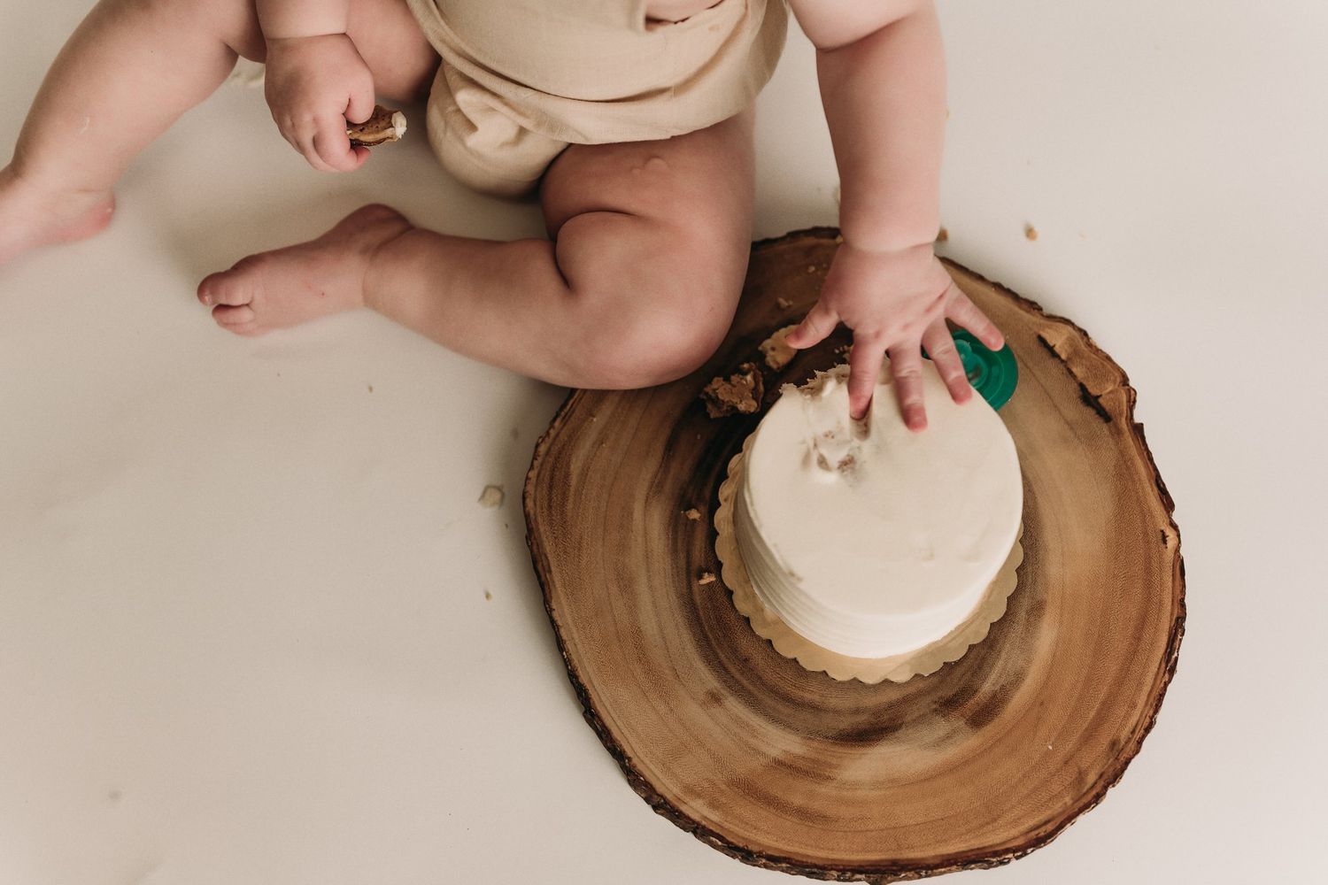 Small hands reach toward white cake placed on rustic wooden slice from above.
